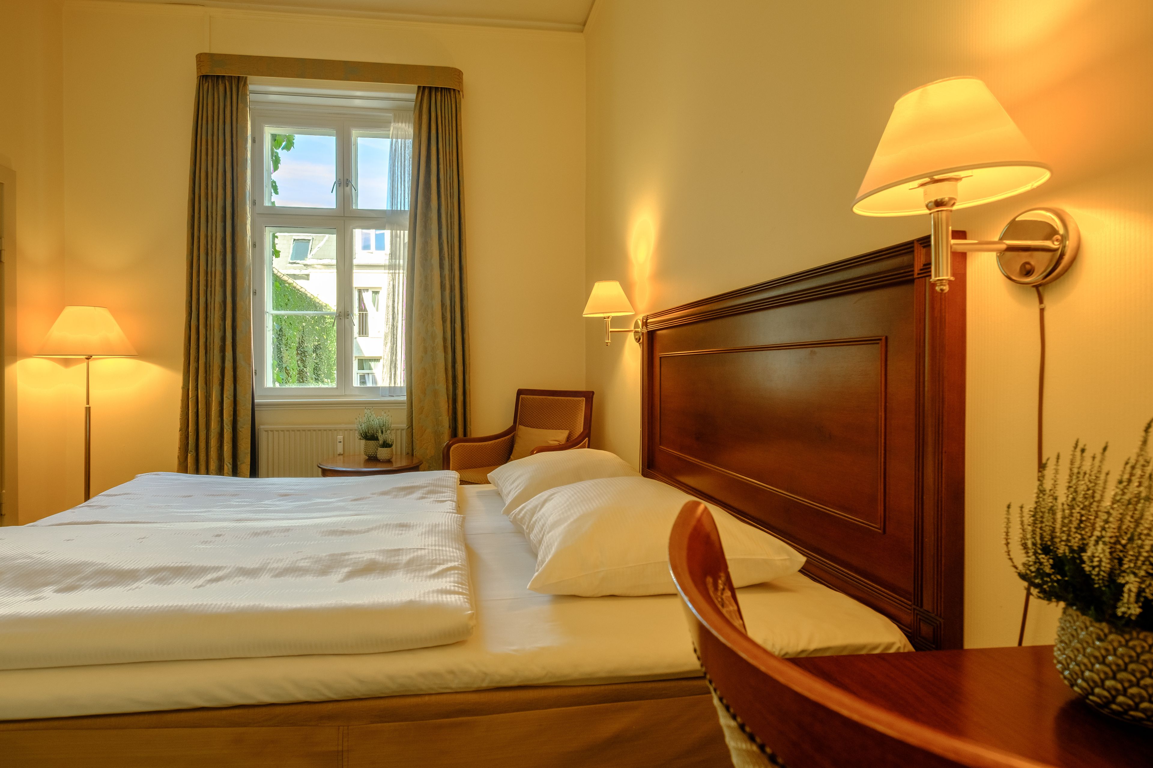 A hotel room with a large bed, two bedside lamps, a wooden headboard, a nightstand, and a window with curtains.