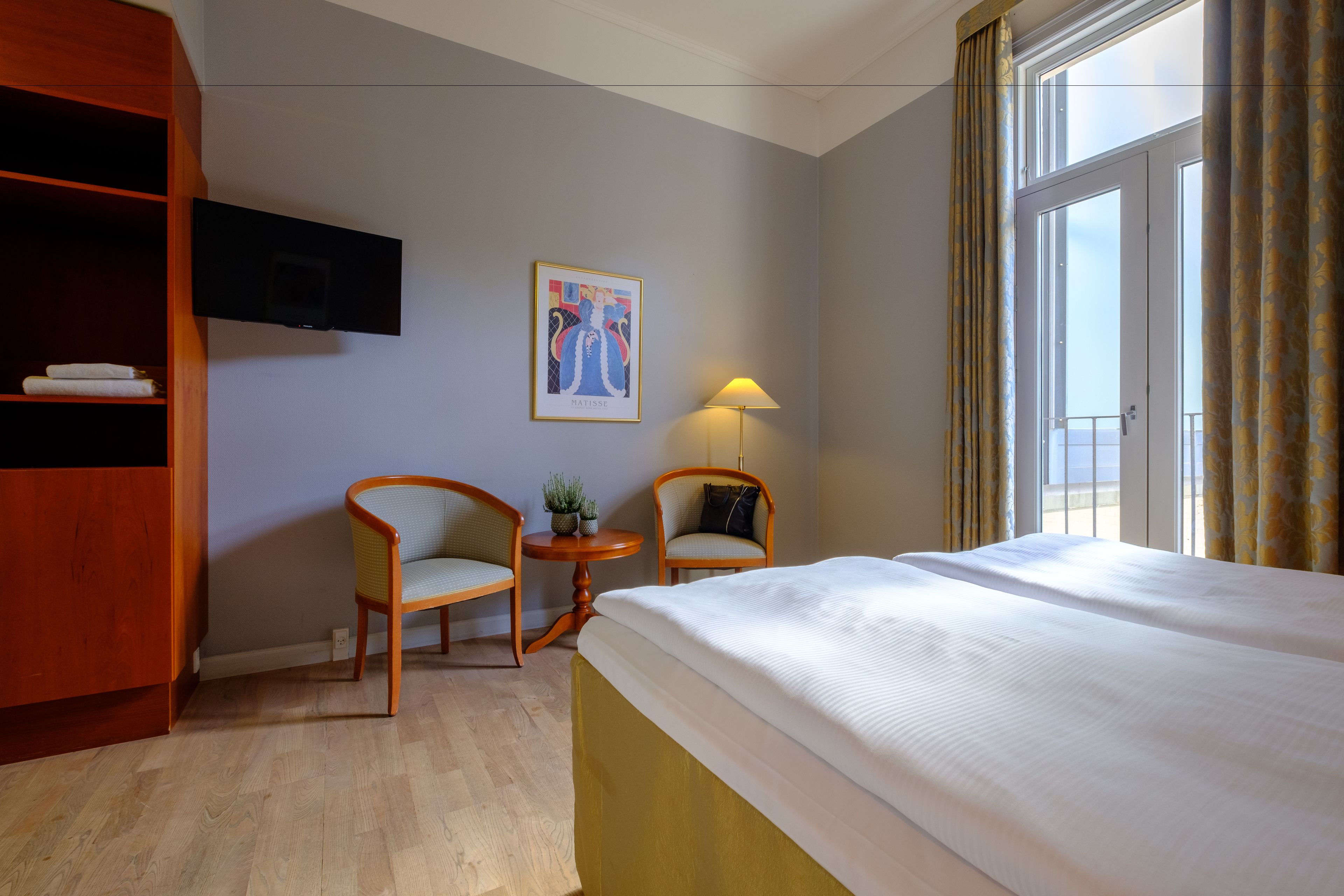 Quarto de hotel com cama, duas cadeiras, uma mesinha com abajur, televisão na parede e janela com cortinas.