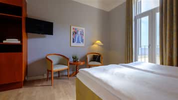 Quarto de hotel com cama, duas cadeiras, uma mesinha com abajur, televisão na parede e janela com cortinas.