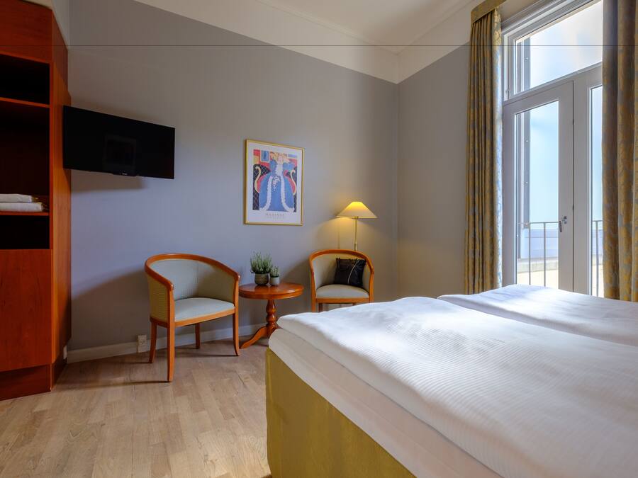 Quarto de hotel com cama, duas cadeiras, uma mesinha com abajur, televisão na parede e janela com cortinas.