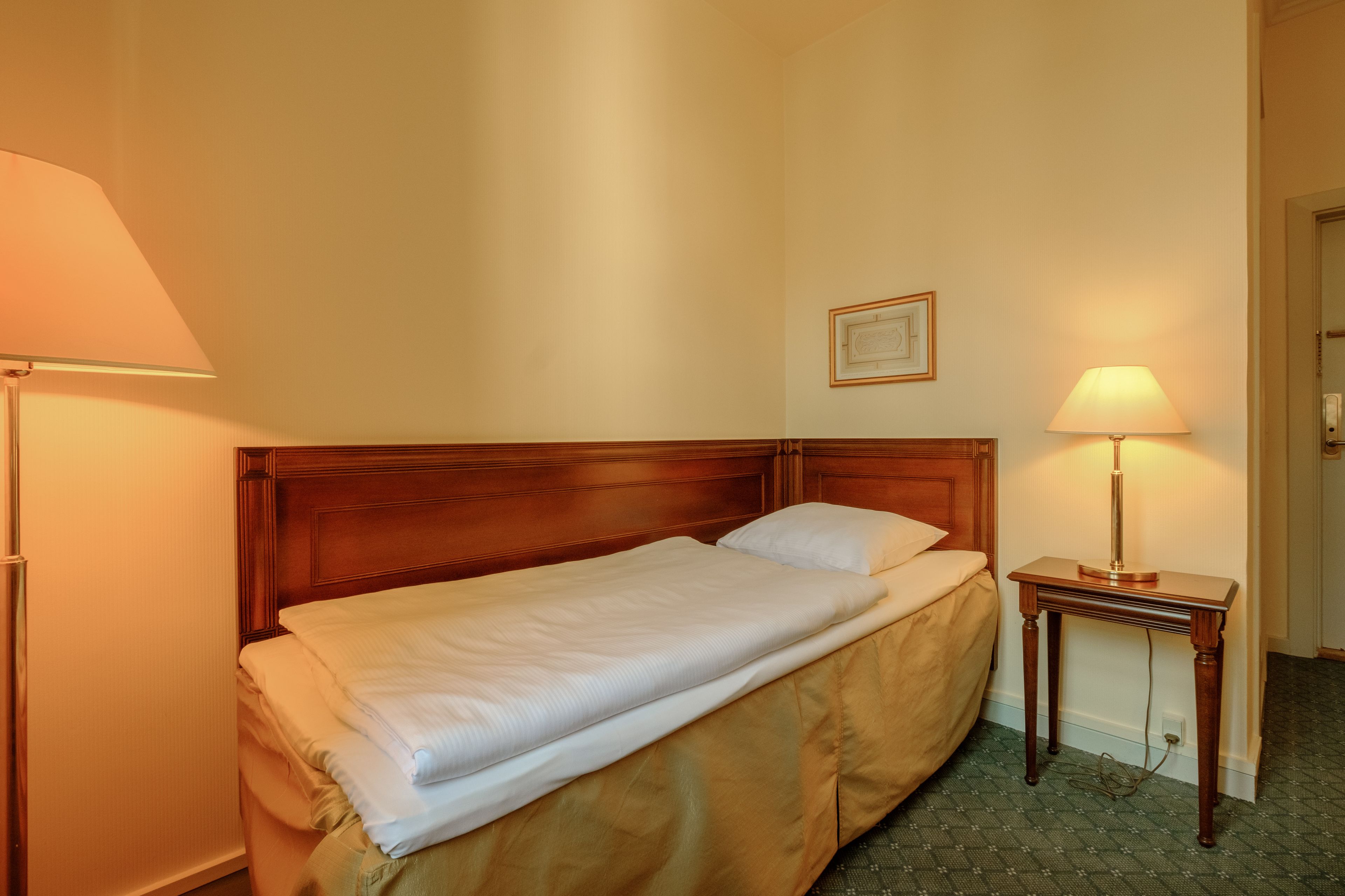 A neatly made bed with a wooden headboard, a bedside table with a lamp, and a framed picture on the wall.