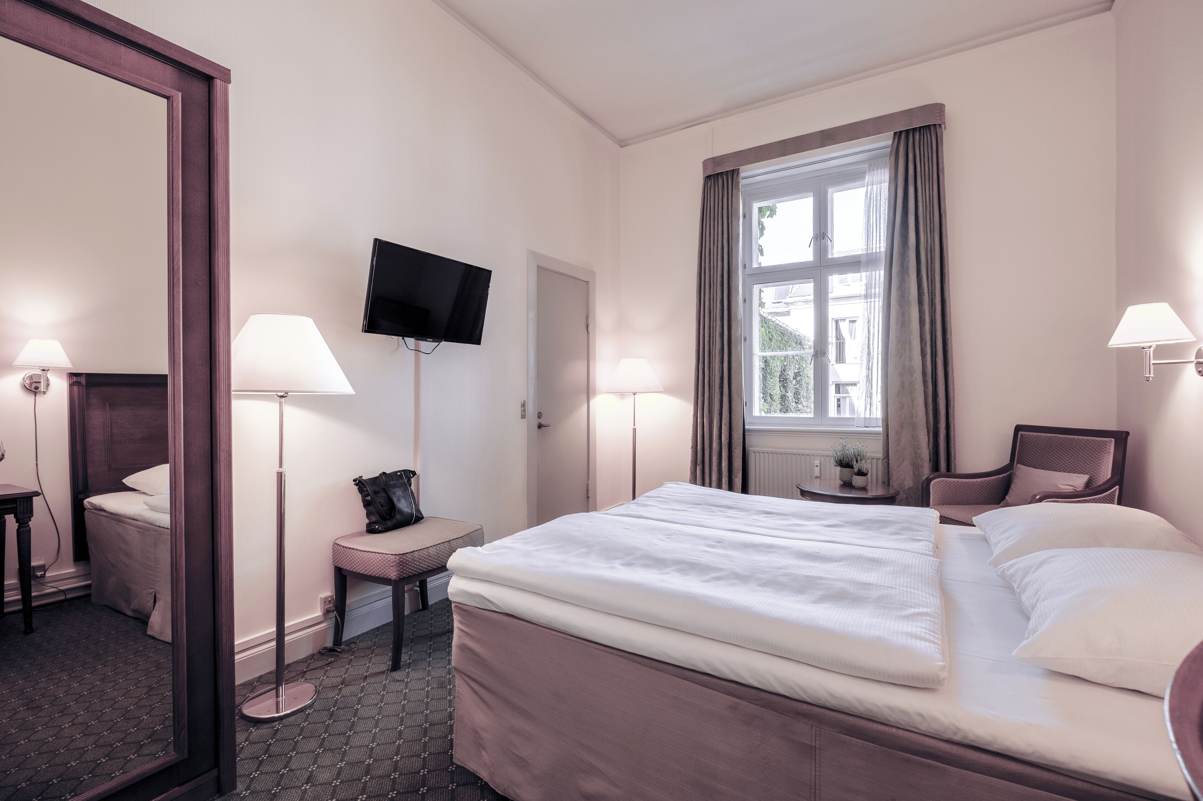 A hotel room with a large bed, two bedside lamps, a flat-screen TV, a window with curtains, a chair, and a small table with a lamp.