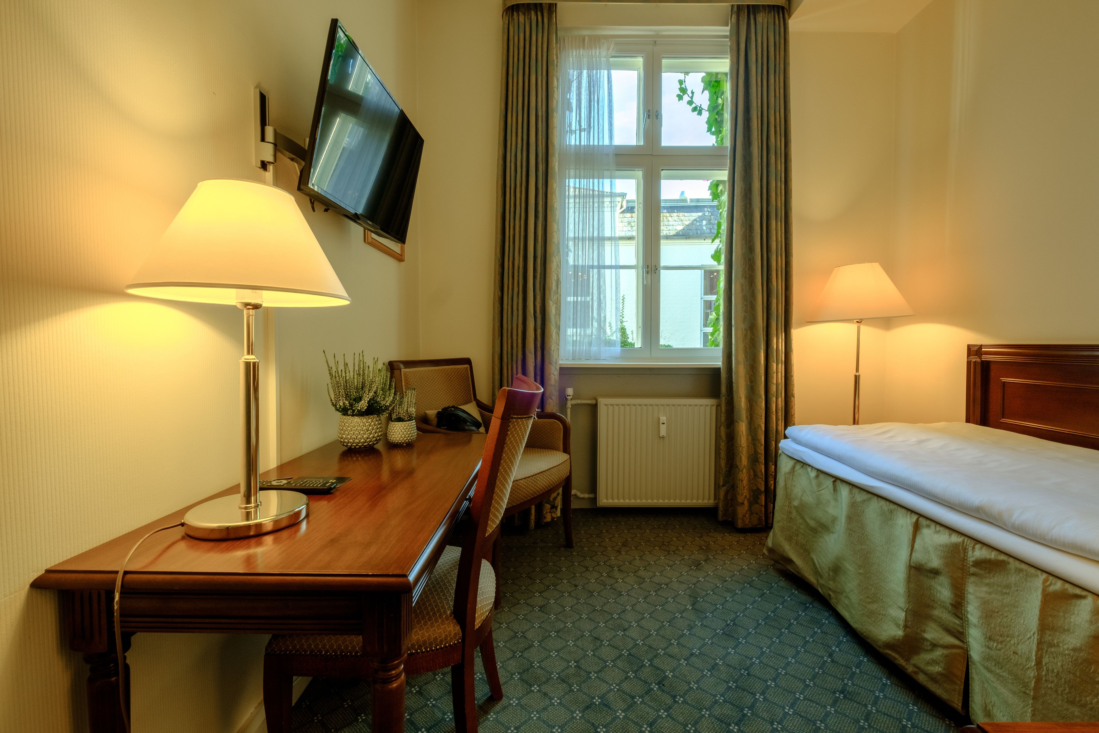 A hotel room with a wooden desk, two chairs, a bed, a TV mounted on the wall, and a window with curtains.