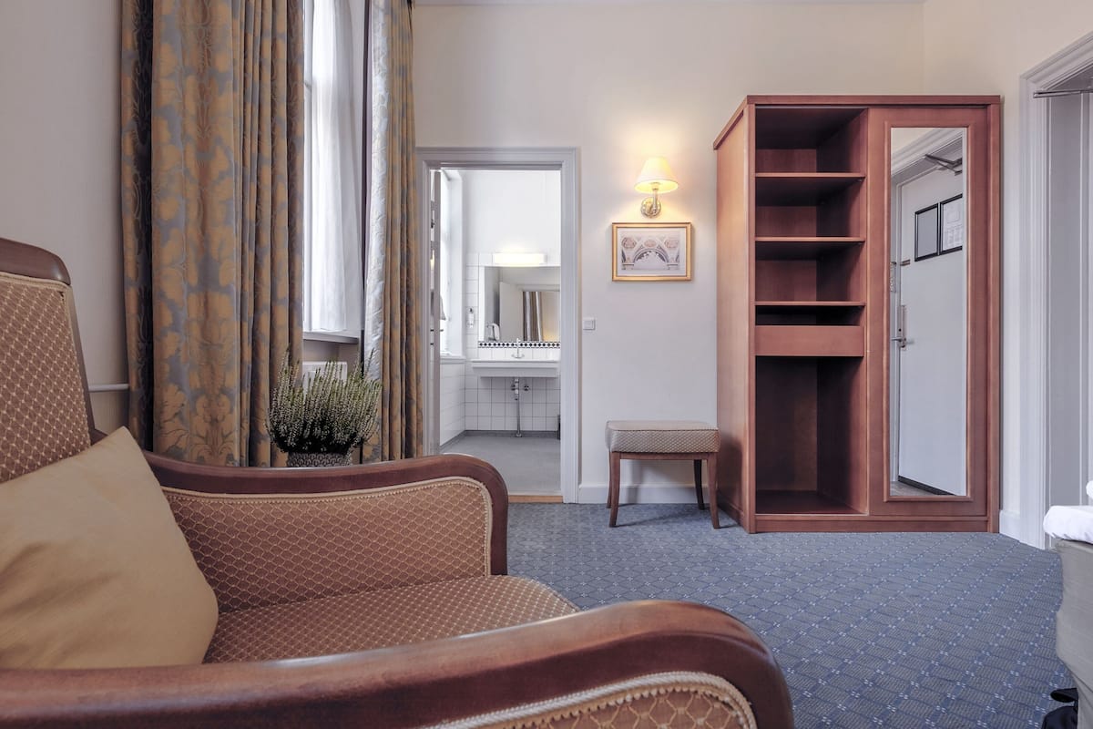 Habitación de hotel con un sillón marrón, un armario de madera, un pequeño banco y un baño visible a través de una puerta abierta.