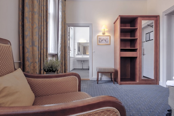 Habitación de hotel con un sillón marrón, un armario de madera, un pequeño banco y un baño visible a través de una puerta abierta.