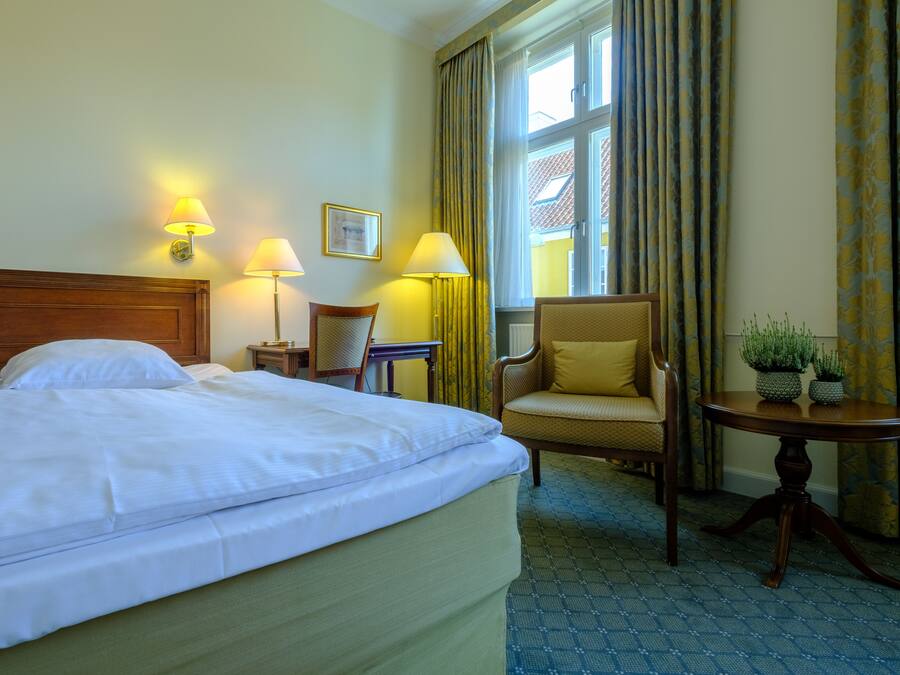 A hotel room with a large bed, two bedside lamps, a chair, a small table with plants, and a window with curtains.
