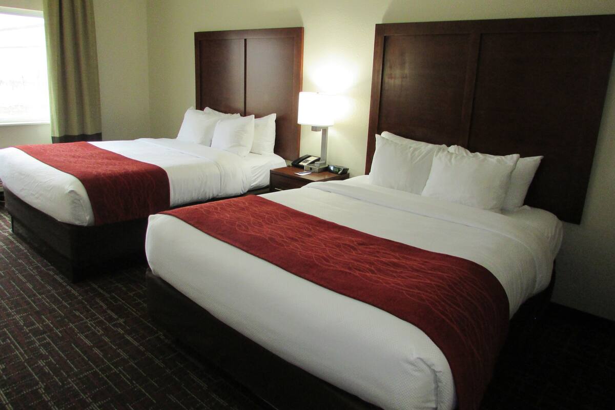 A hotel room with two beds, each with a wooden headboard, white bedding, and a red accent. There is a nightstand with a lamp and a telephone.
