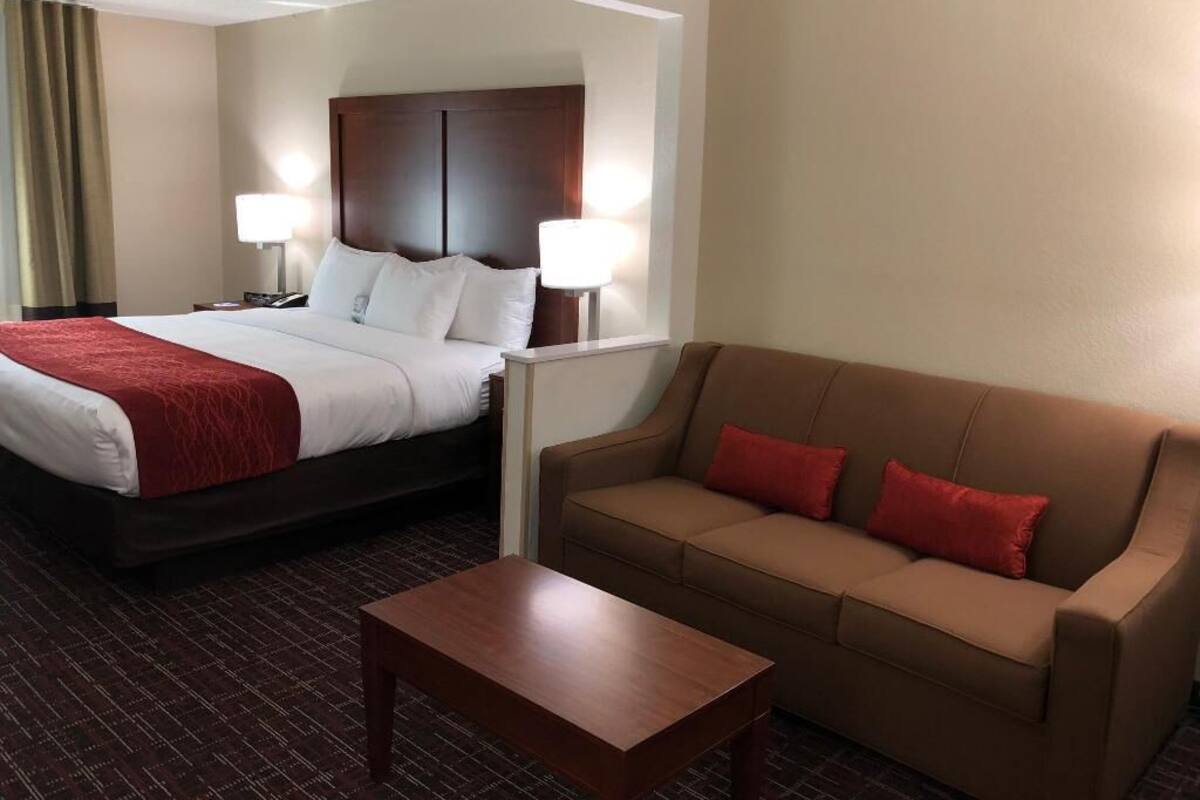 A hotel room with a bed, two bedside lamps, a brown sofa with red cushions, and a wooden coffee table.