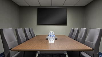A conference room with a long wooden table, six grey upholstered chairs, and a large blank screen on the wall.