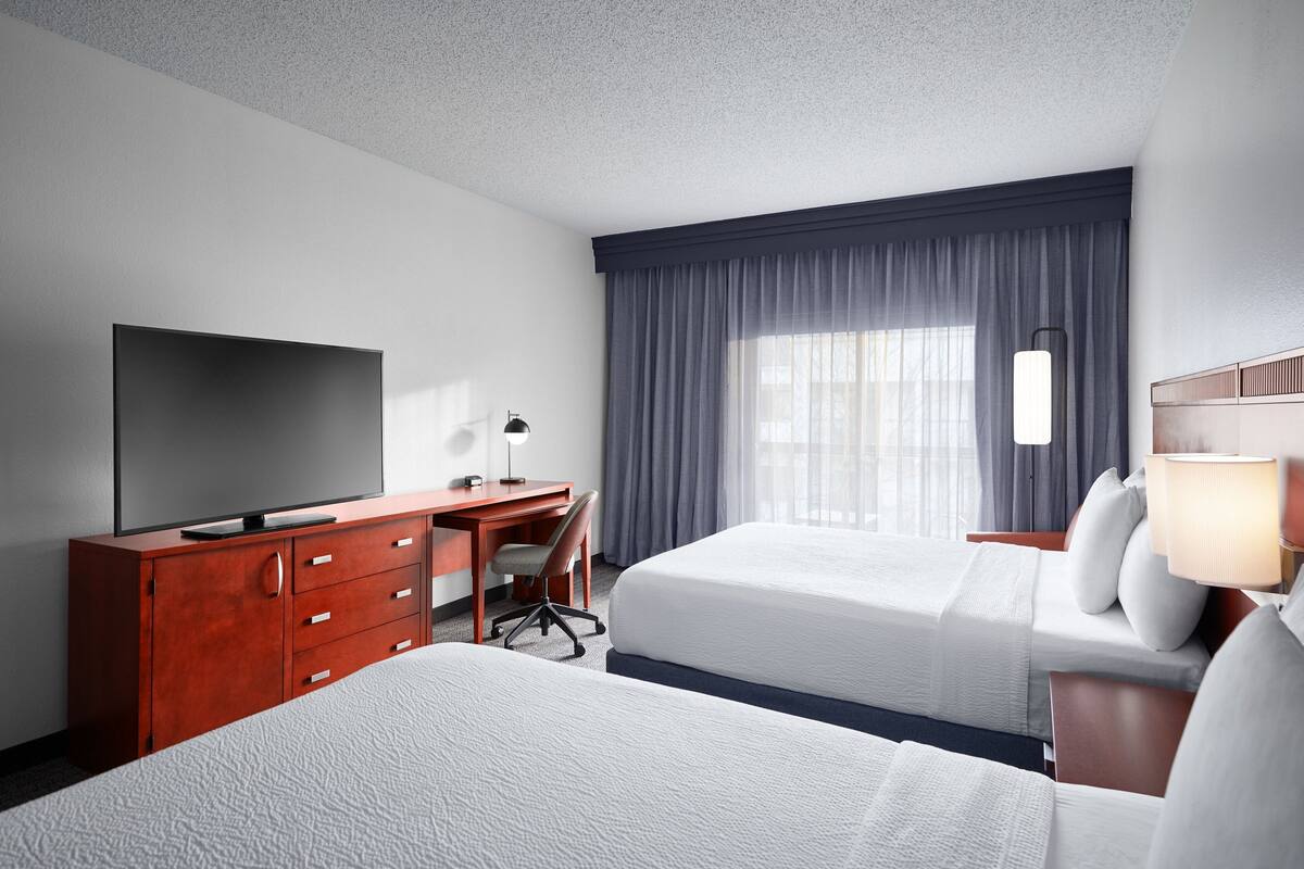 Ein Hotelzimmer mit zwei Betten, einem Schreibtisch mit Fernseher und einem Fenster mit Vorhängen.