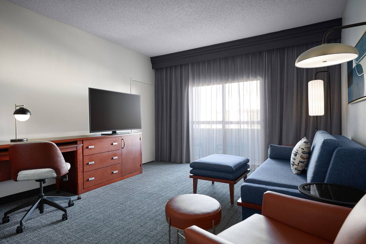 Ein Hotelzimmer mit Flachbildfernseher, Schreibtisch mit Schubladen, blauem Sofa, rundem Hocker und großem Fenster mit Vorhängen.