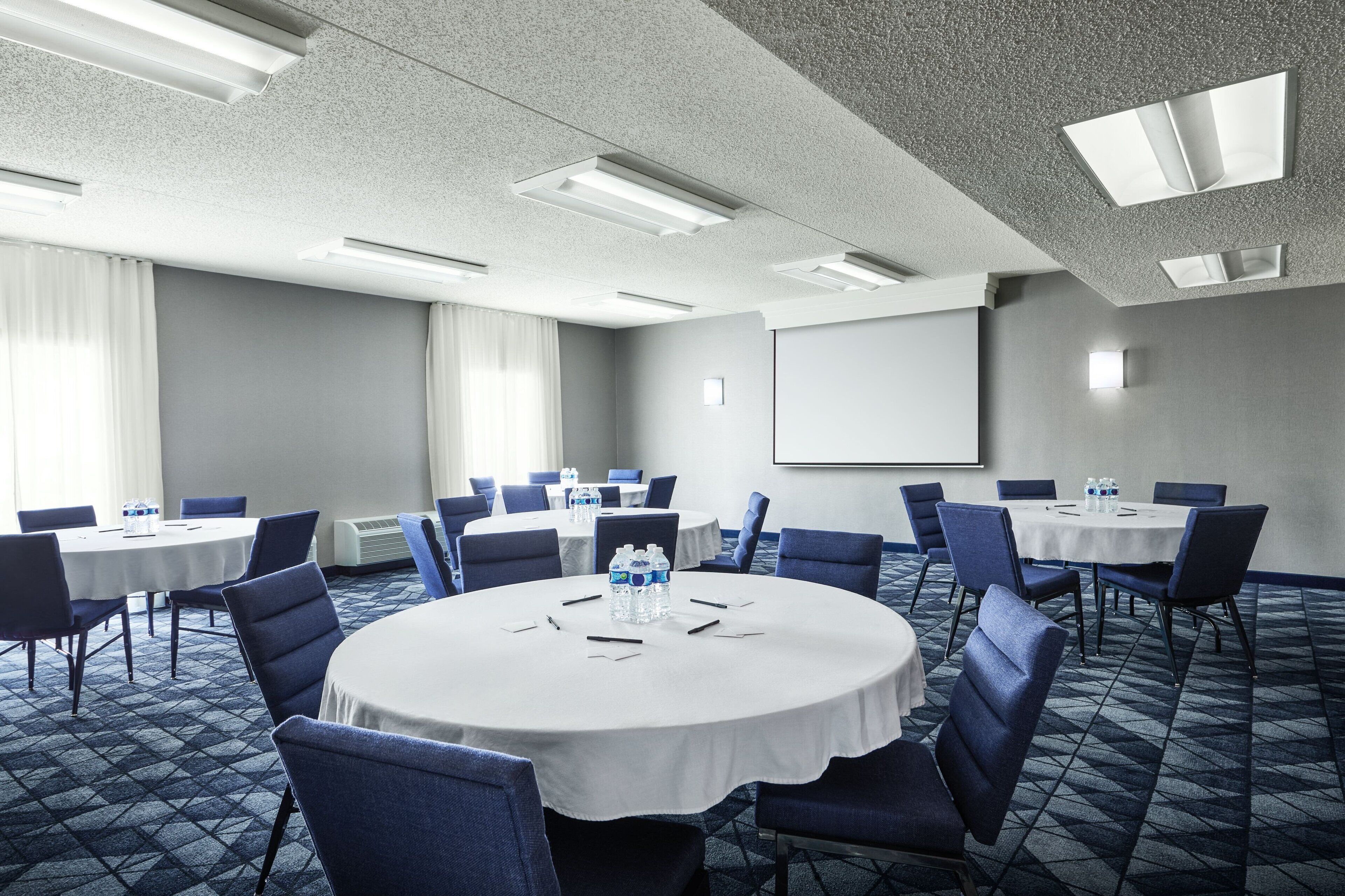A conference room with round tables, blue chairs, and a projector screen on the wall.