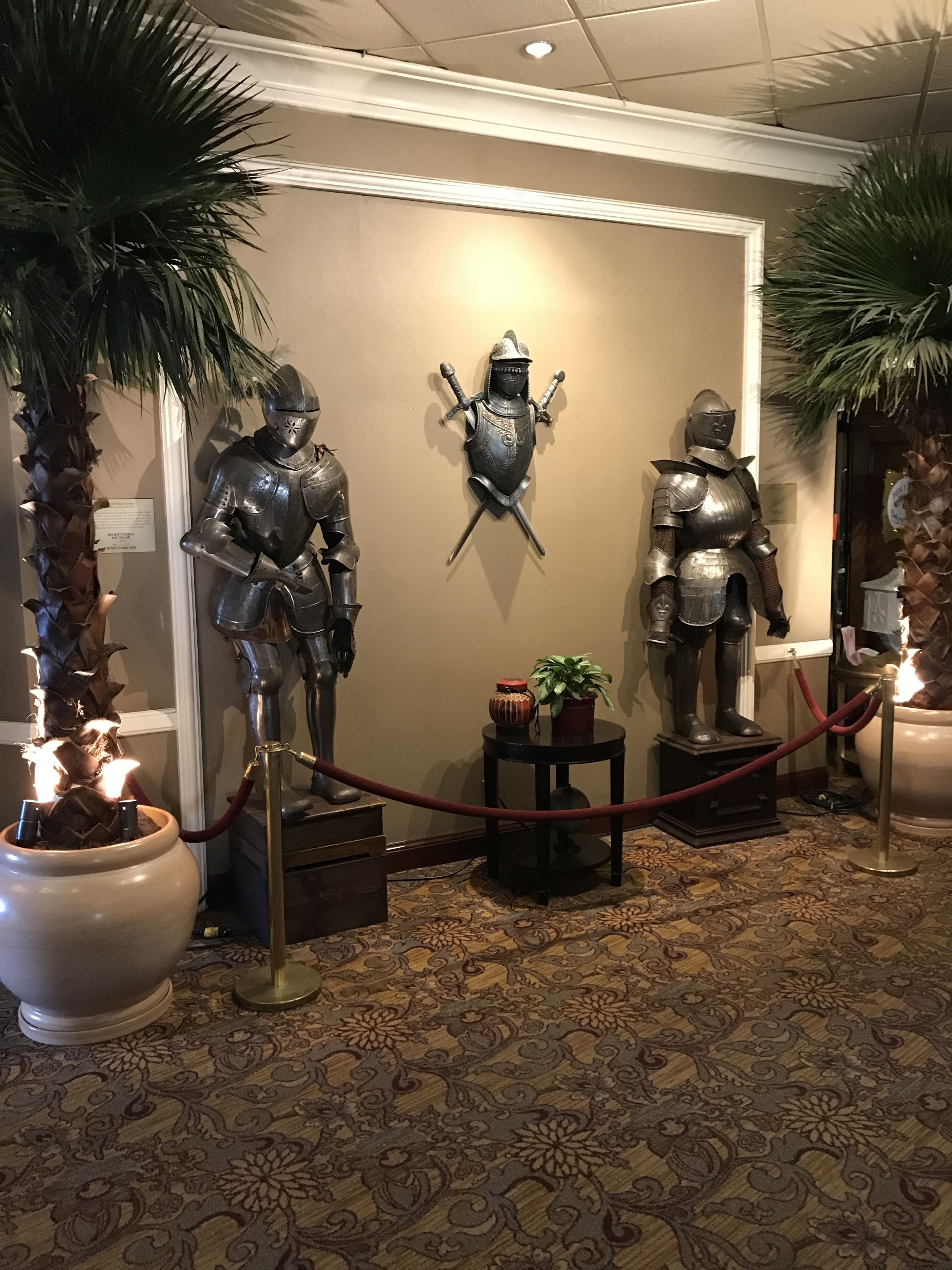 A display of medieval armor and a shield with swords, surrounded by potted plants and a small table with a vase.