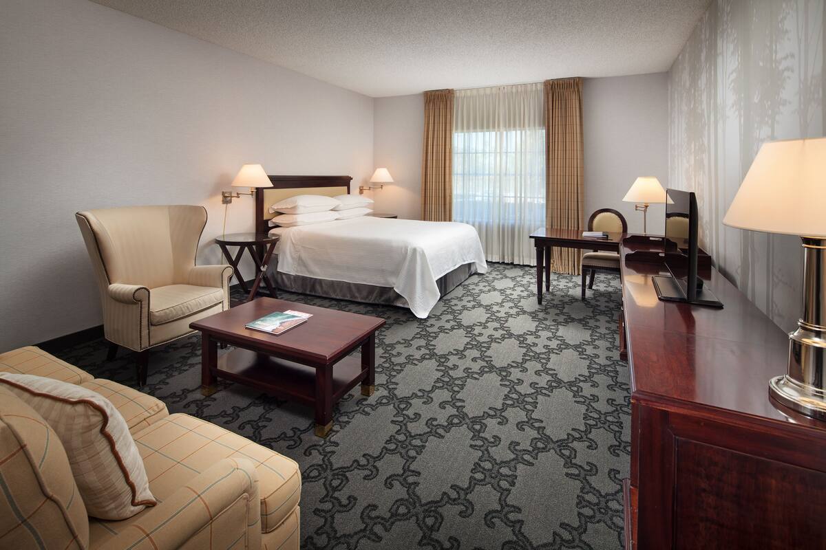 A hotel room with a bed, two bedside tables with lamps, a chair, a sofa, and a desk with a lamp.
