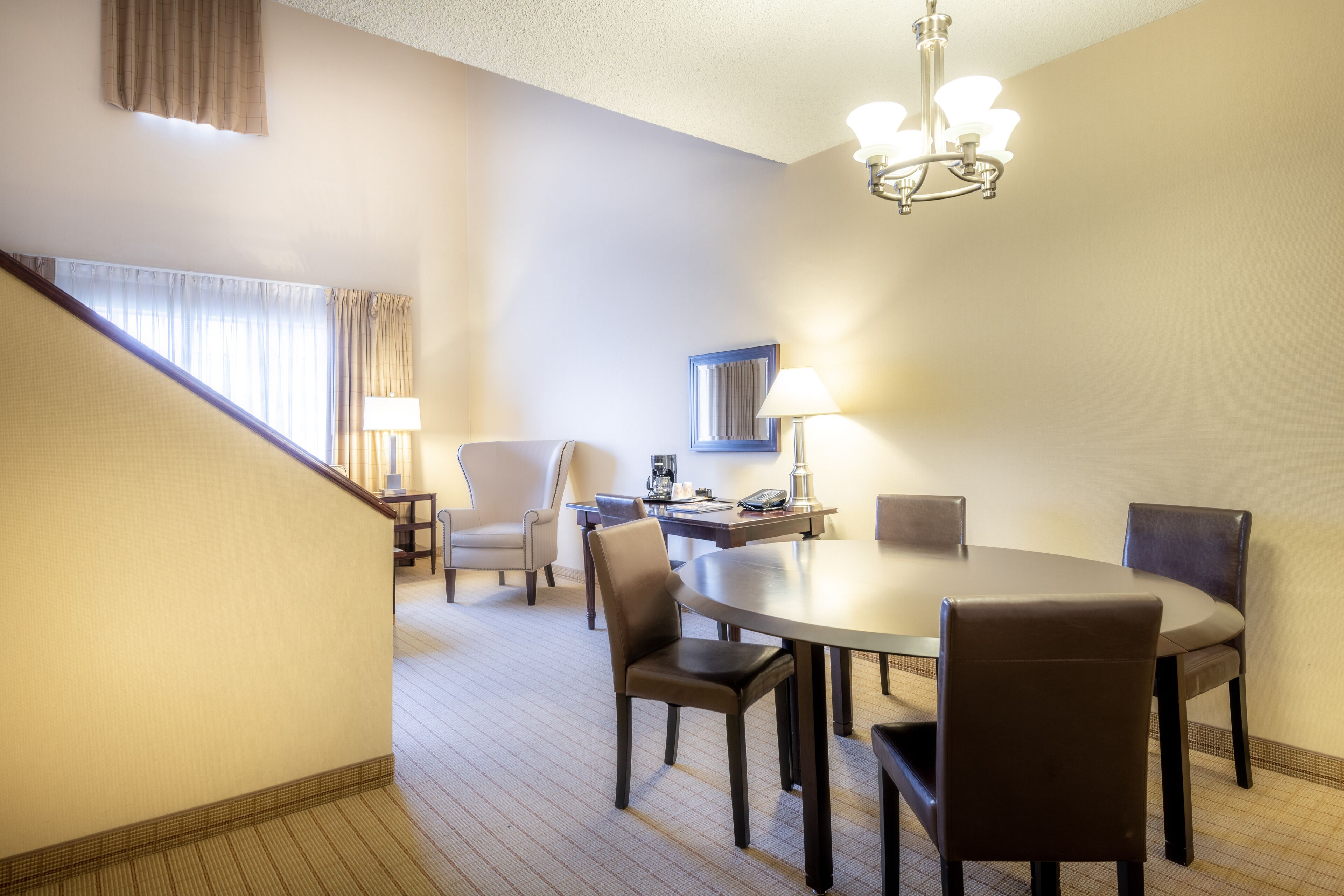 A hotel dining area with a round table, six chairs, a chandelier, a lamp, a coffee maker, and a framed picture on the wall.