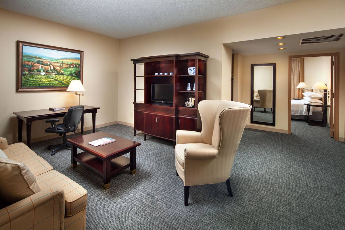A hotel room with a sofa, a desk with a lamp, a television, a cabinet, and a chair.