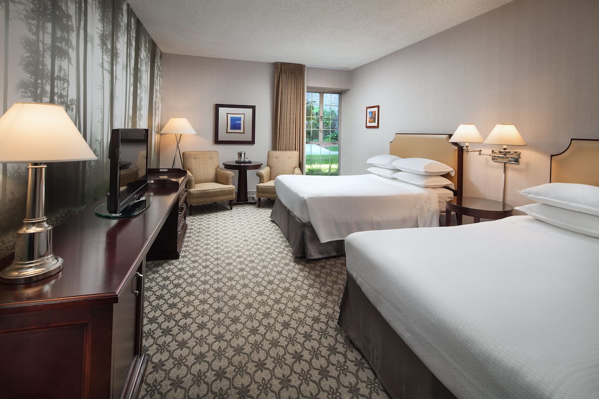 A hotel room with two beds, a desk with a TV, a chair, a small table, and a window with a view of greenery.