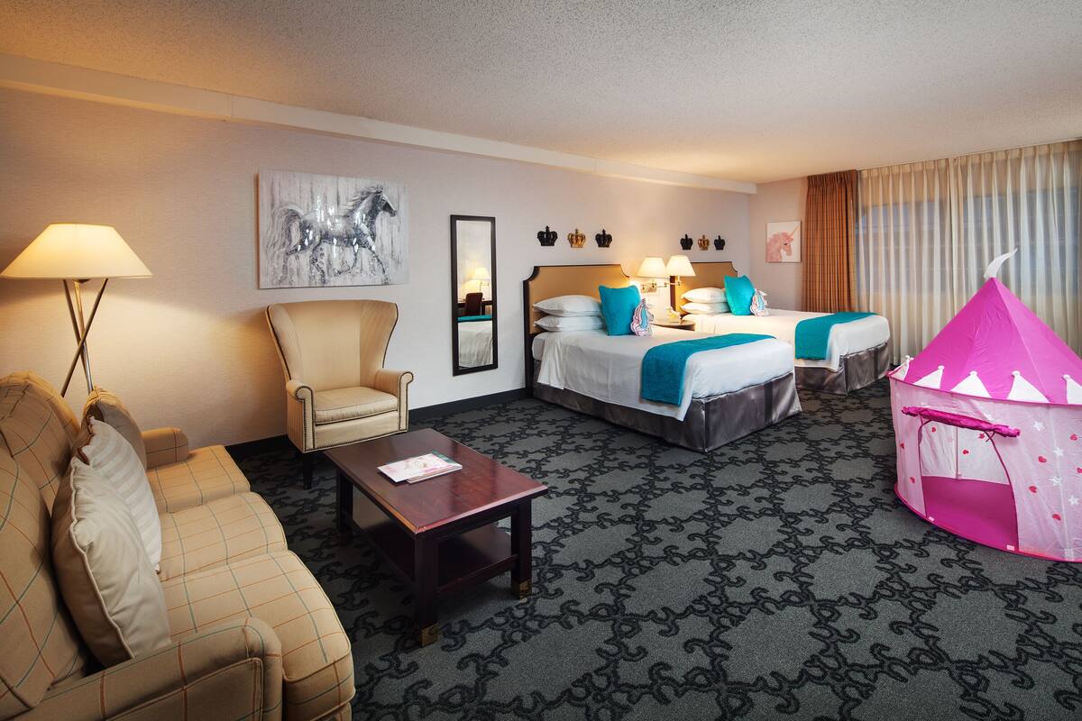 A hotel room with a sofa, two beds, a chair, a small table, and a pink play tent.