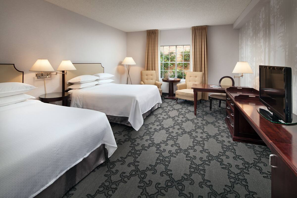 A hotel room with two beds, a desk with a TV, a chair, and a small table.