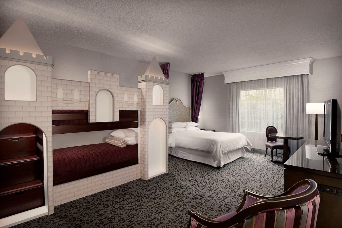 A hotel room with a castle-themed bunk bed, a separate bed, a desk with a chair, and a television.