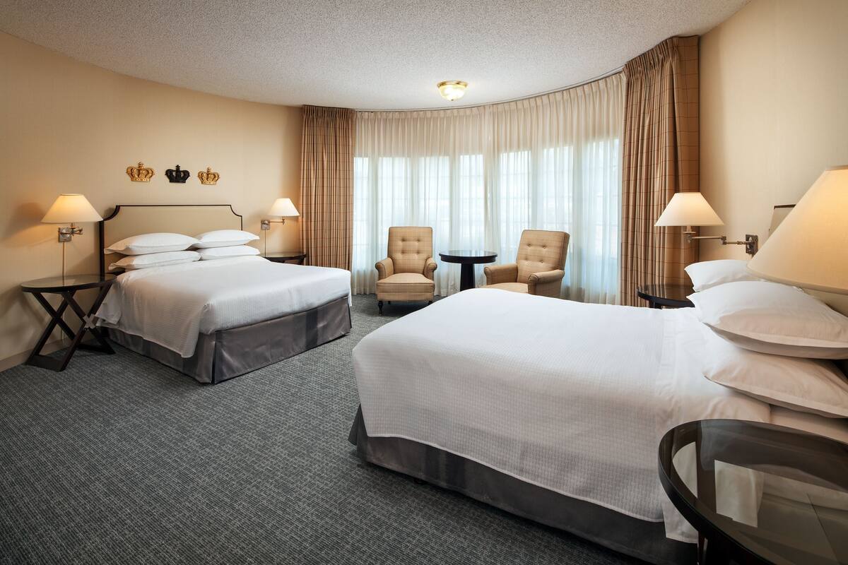 A hotel room with two beds, a small table, two armchairs, and a large window with curtains.