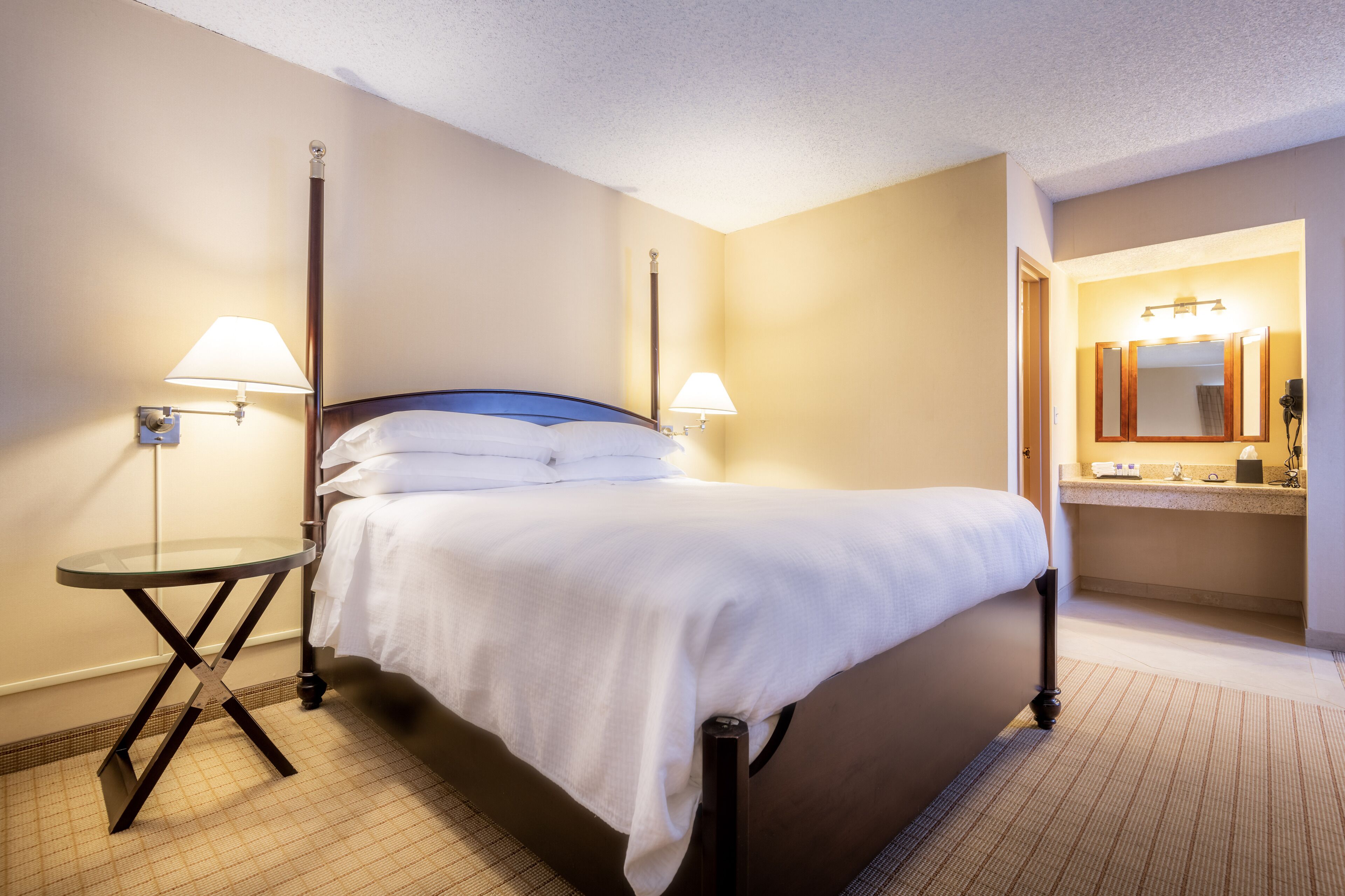 A hotel room with a bed, two bedside lamps, a small round table, and a bathroom with a mirror and sink.
