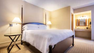 A hotel room with a bed, two bedside lamps, a small round table, and a bathroom with a mirror and sink.
