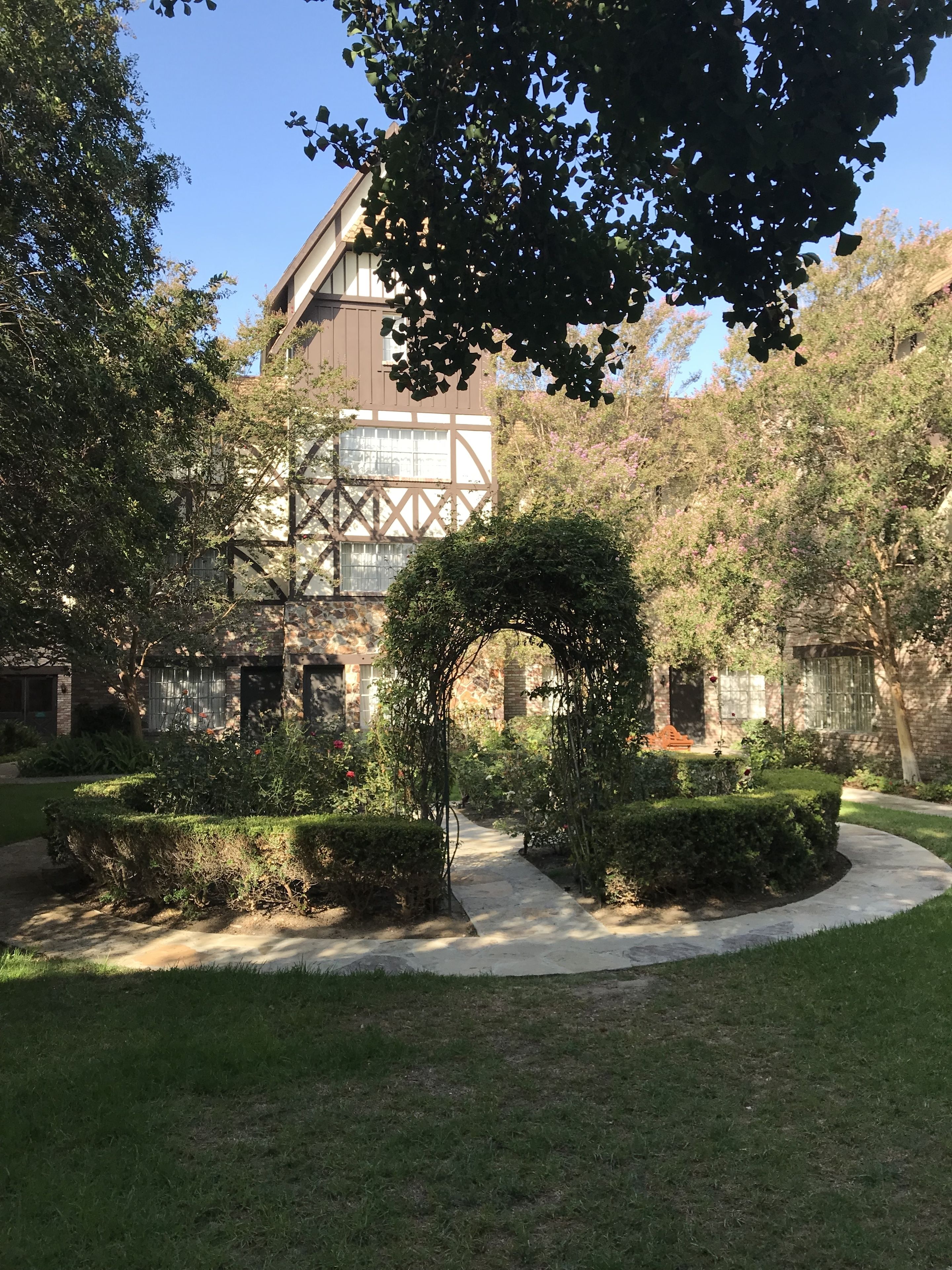 Ein Innenhof mit einem gepflasterten Weg, ordentlich geschnittenen Hecken und einem traditionellen Gebäude mit freiliegenden Holzbalken und großen Fenstern.