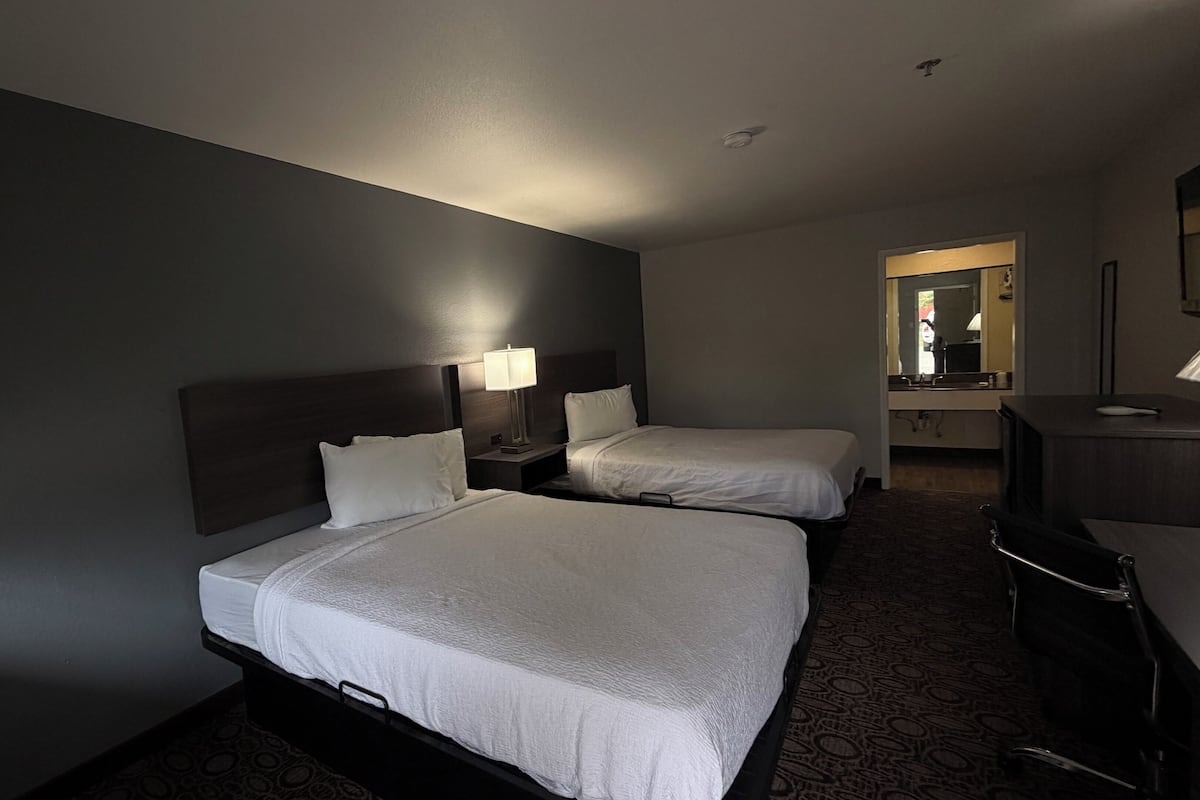 A hotel room with two beds, a desk, a chair, a mirror, and a lamp.