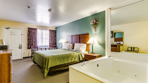 Ein Hotelzimmer mit Bett, Schreibtisch, Stuhl, Nachttisch mit Lampe sowie einem Badezimmer mit Badewanne und Spiegel.