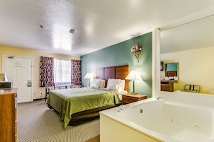 Habitación de hotel con cama, escritorio, silla, mesita de noche con lámpara, baño con bañera y espejo.
