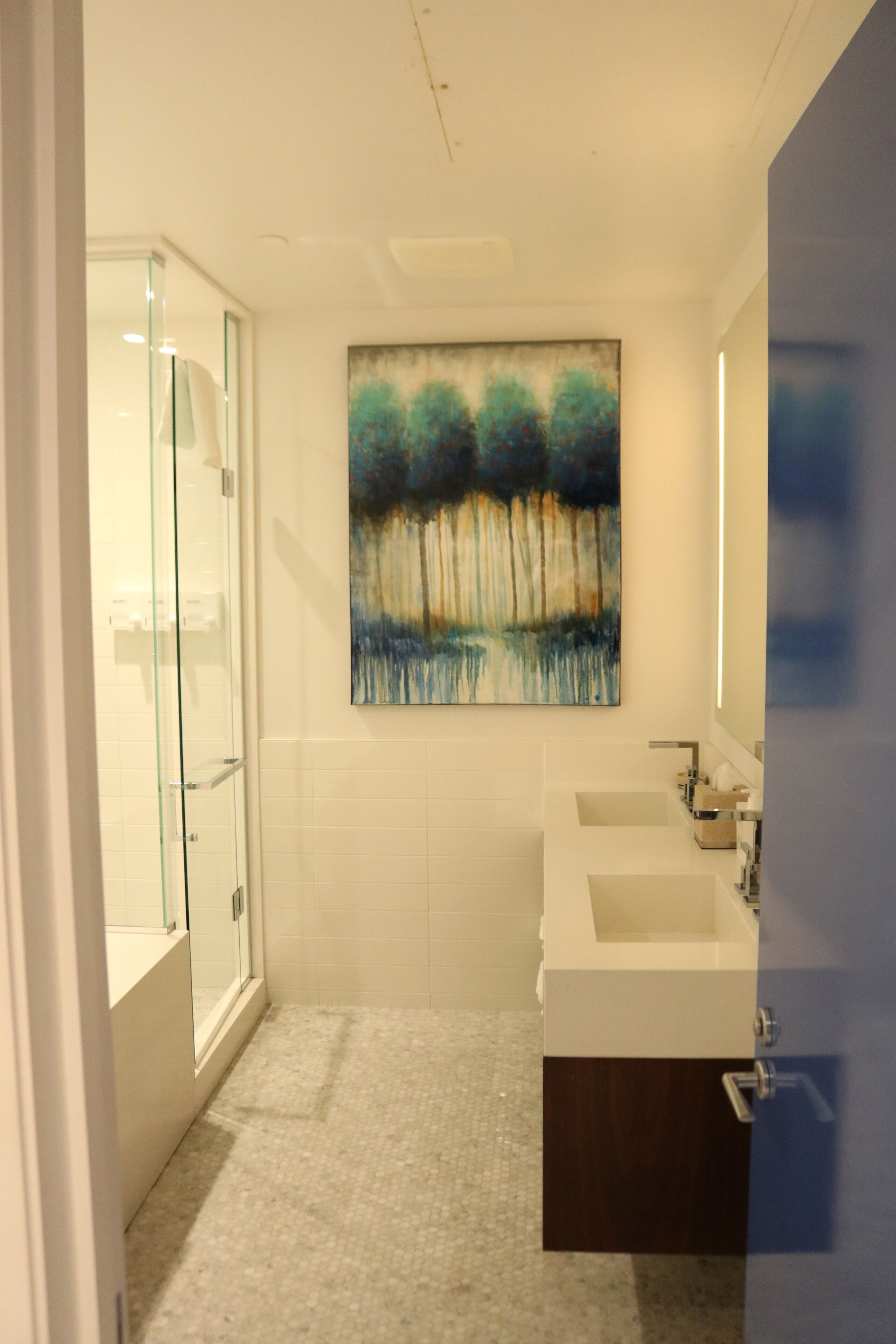 A bathroom with a glass shower enclosure, a white sink, and a large abstract painting on the wall.