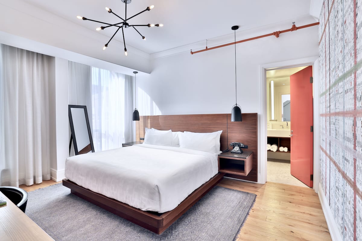 A modern hotel room with a large bed, a wooden headboard, a desk with a chair, a nightstand, and a hanging light fixture.