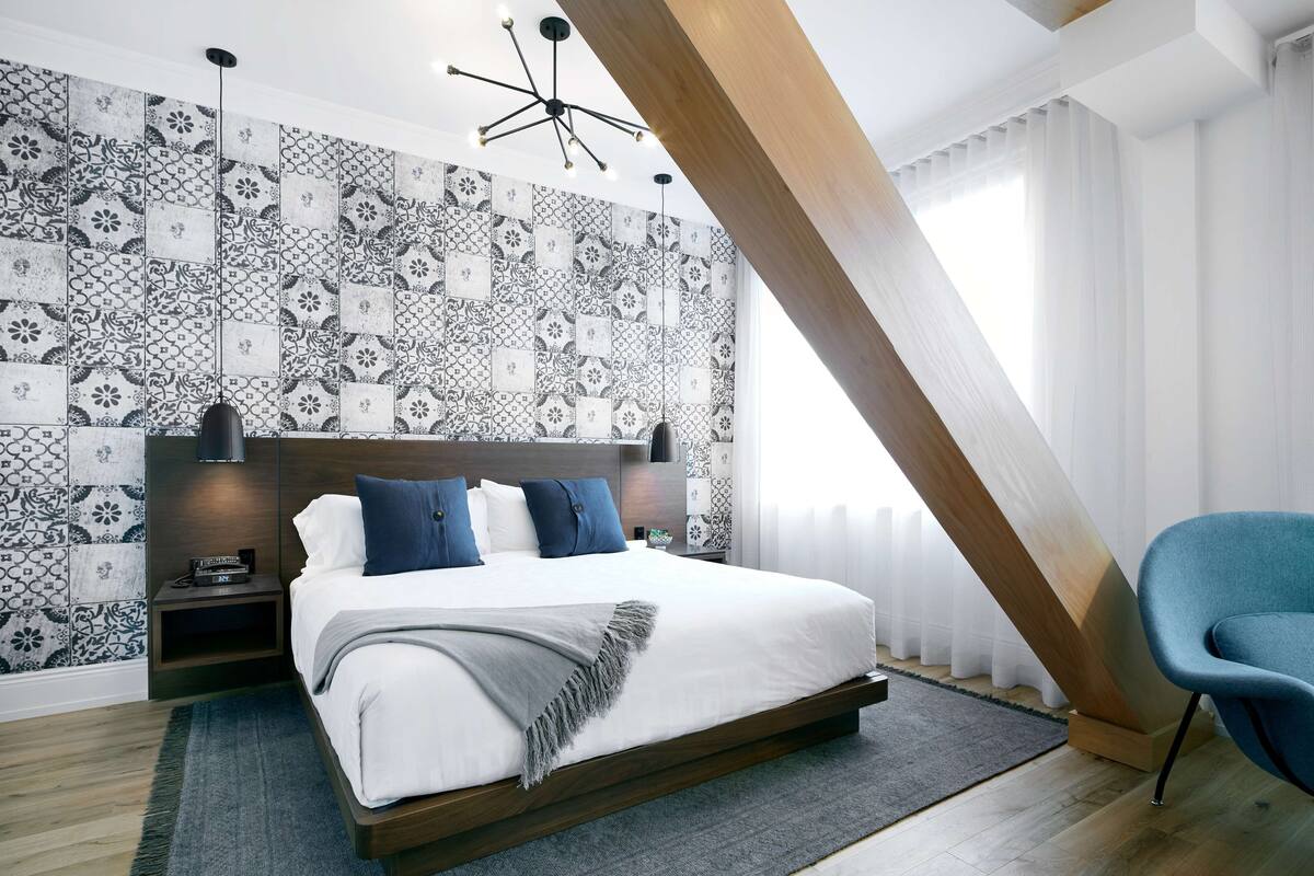 A modern bedroom with a patterned wall, a wooden ceiling beam, a bed with white bedding, a blue chair, and a nightstand with a phone.