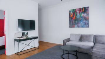 Una sala de estar moderna con un sofá gris, una mesita redonda pequeña, un mueble de televisión negro con televisor de pantalla plana, un cuadro abstracto colorido y piso de madera.