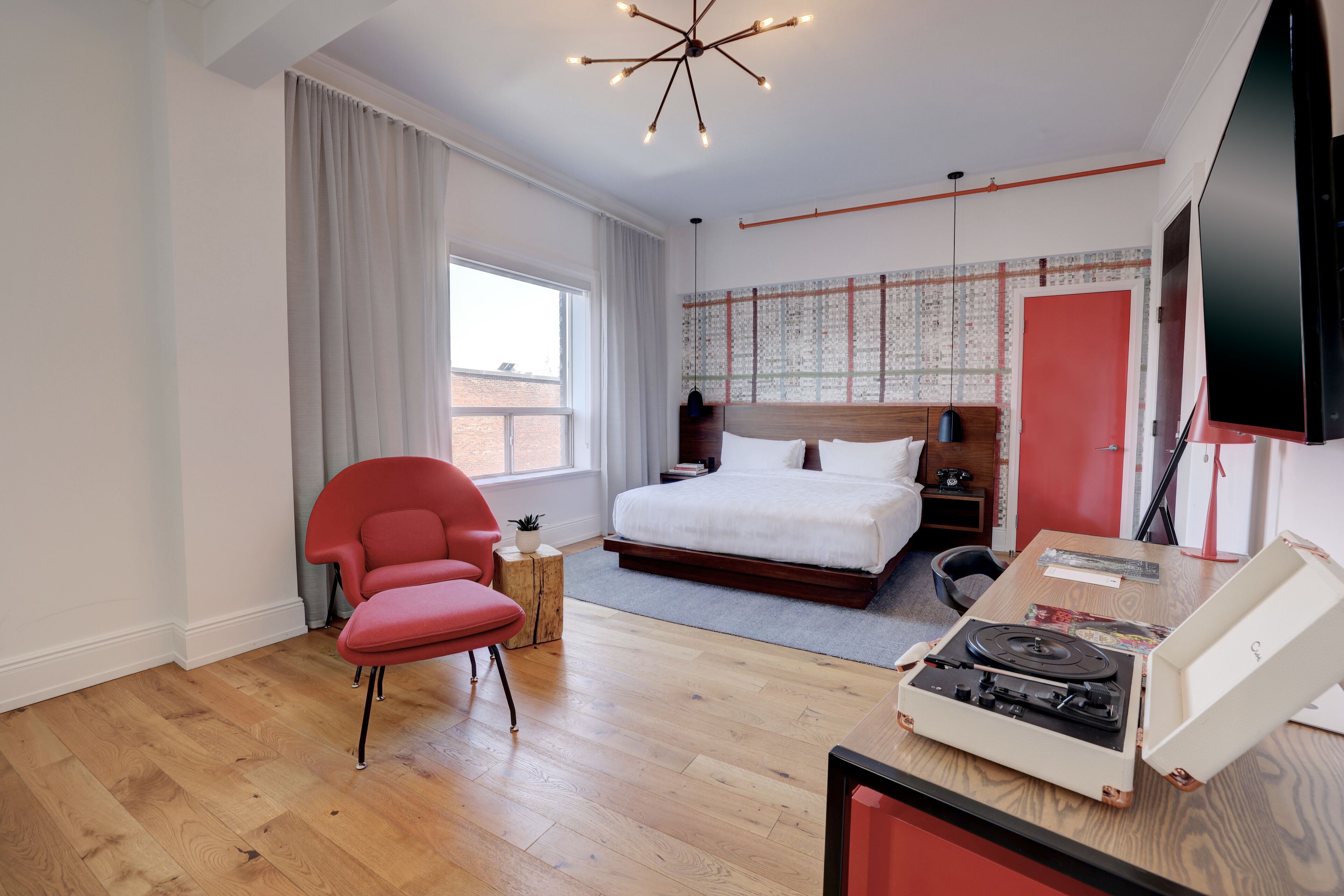 A modern bedroom with a bed, a red chair, a wooden floor, a ceiling light, and a flat-screen TV.