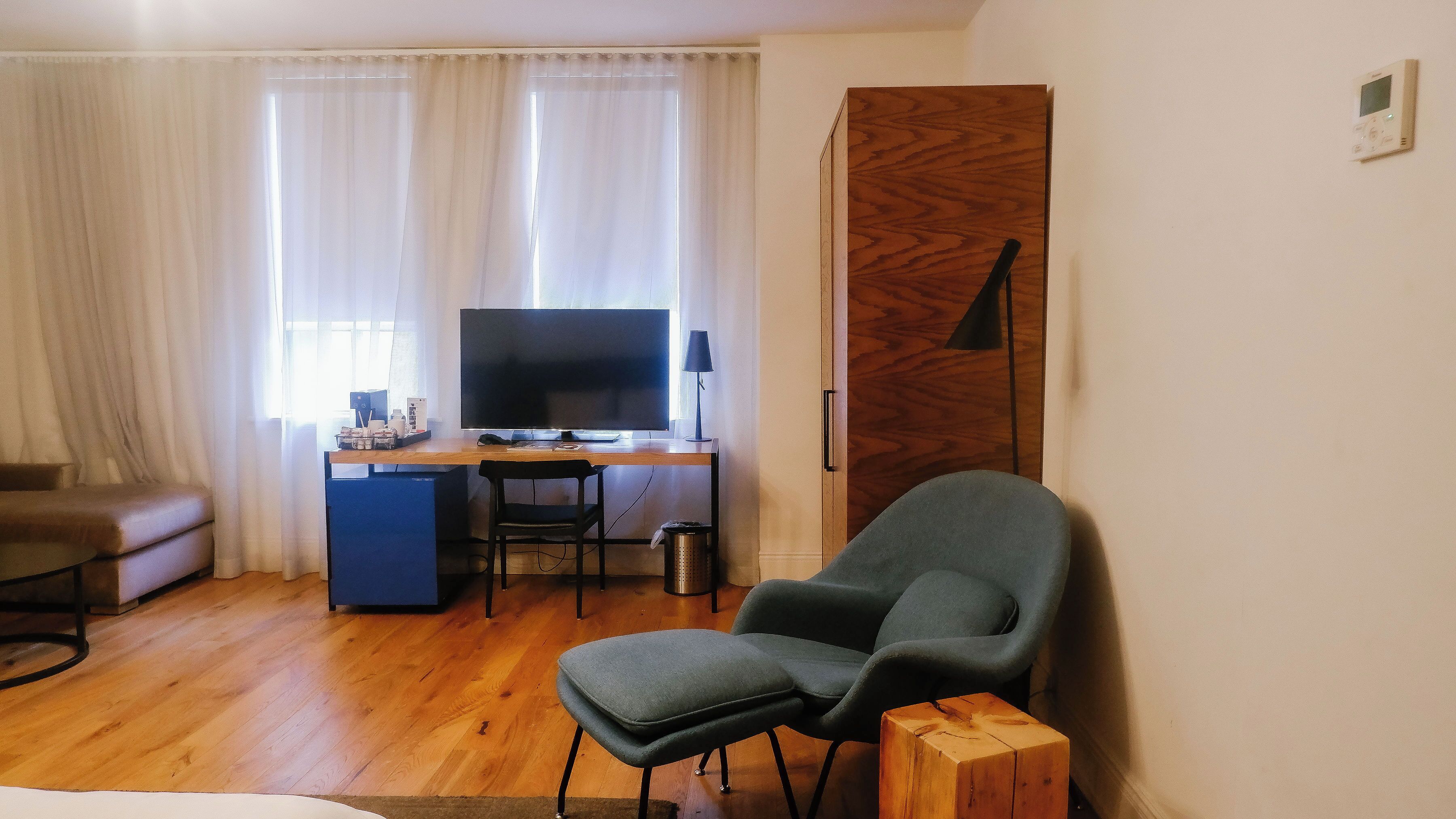 A room with a wooden desk, a large flat-screen TV, a grey armchair, a sofa, and a wooden door.