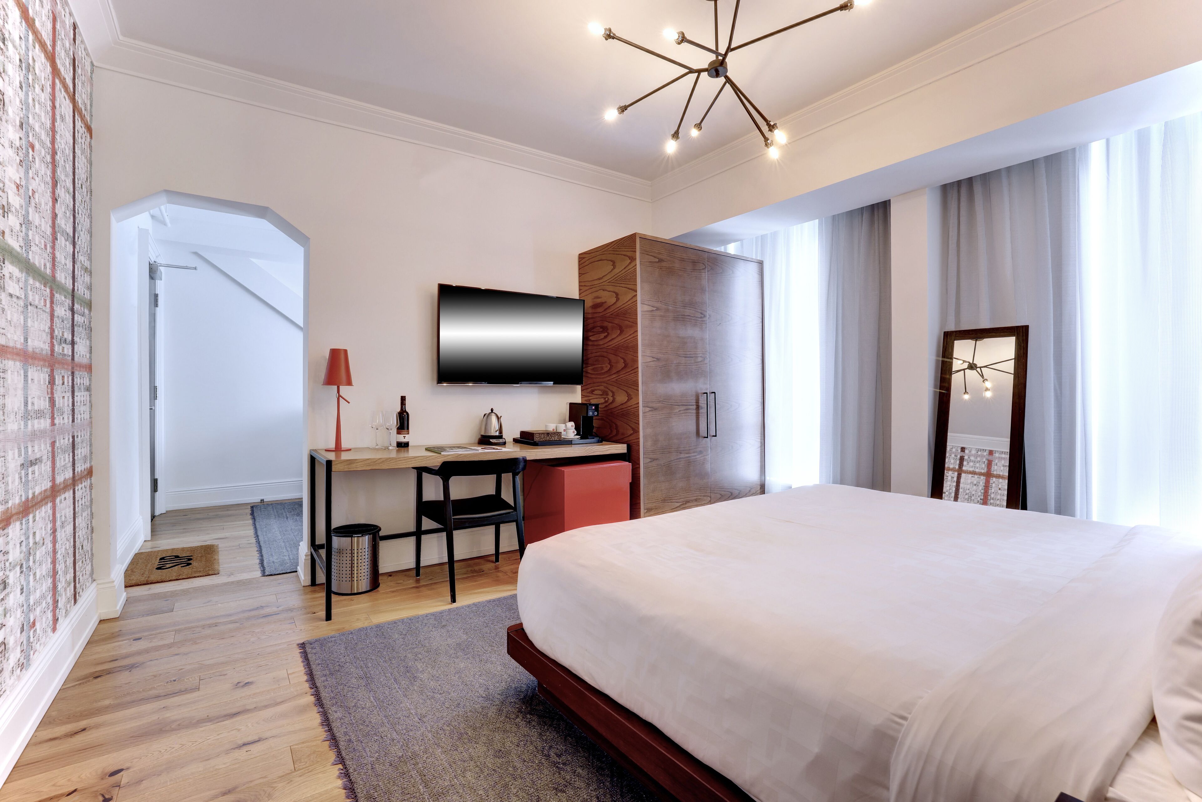 A modern hotel room with a large bed, a desk with a chair, a television, and a wardrobe.