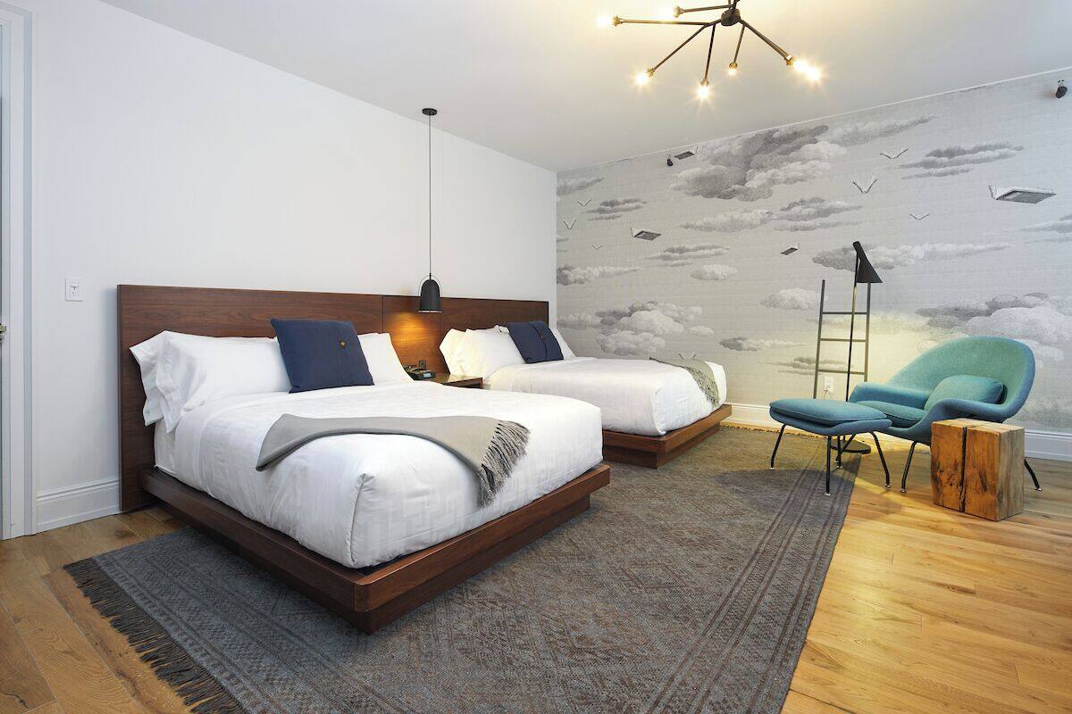 A bedroom with two beds, a nightstand, a chair, and a mural of clouds and birds on the wall.