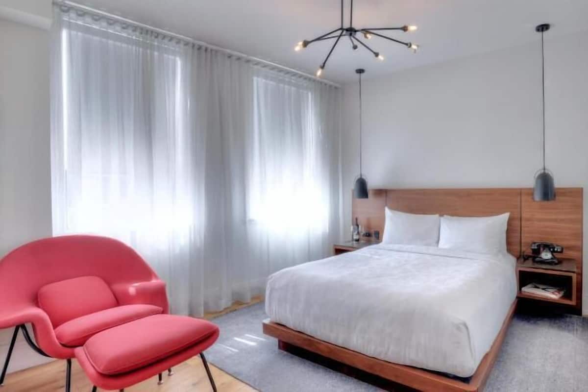 A modern hotel room with a large bed, a red chair, a wooden headboard, and a ceiling light fixture.