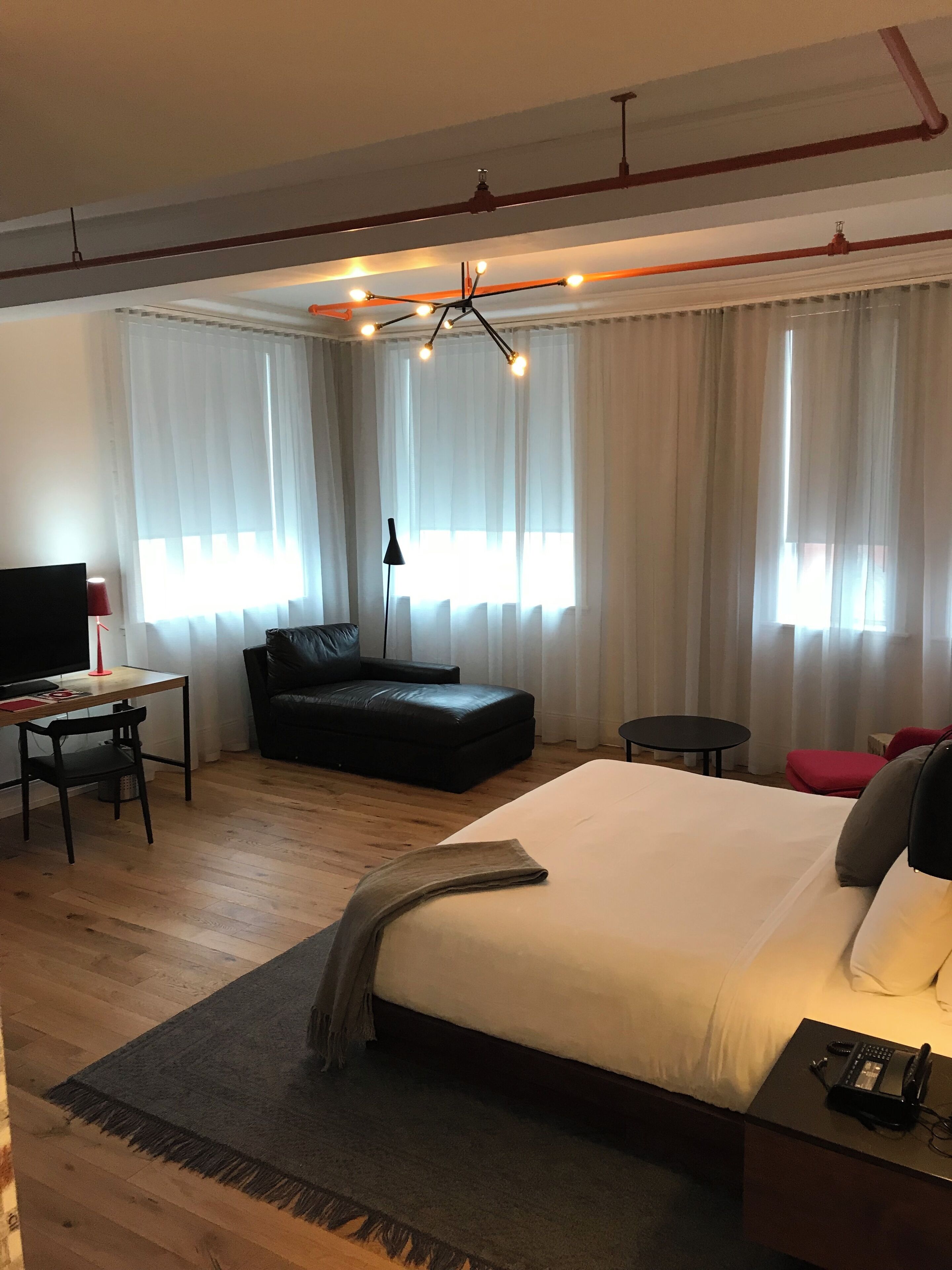 A modern hotel room with a large bed, a desk with a chair, a black leather sofa, and a small table.