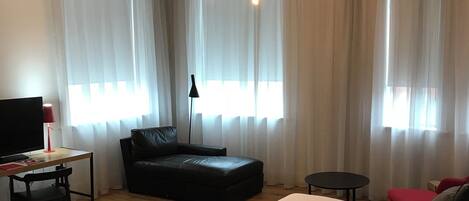 A modern hotel room with a large bed, a desk with a chair, a black leather sofa, and a small table.