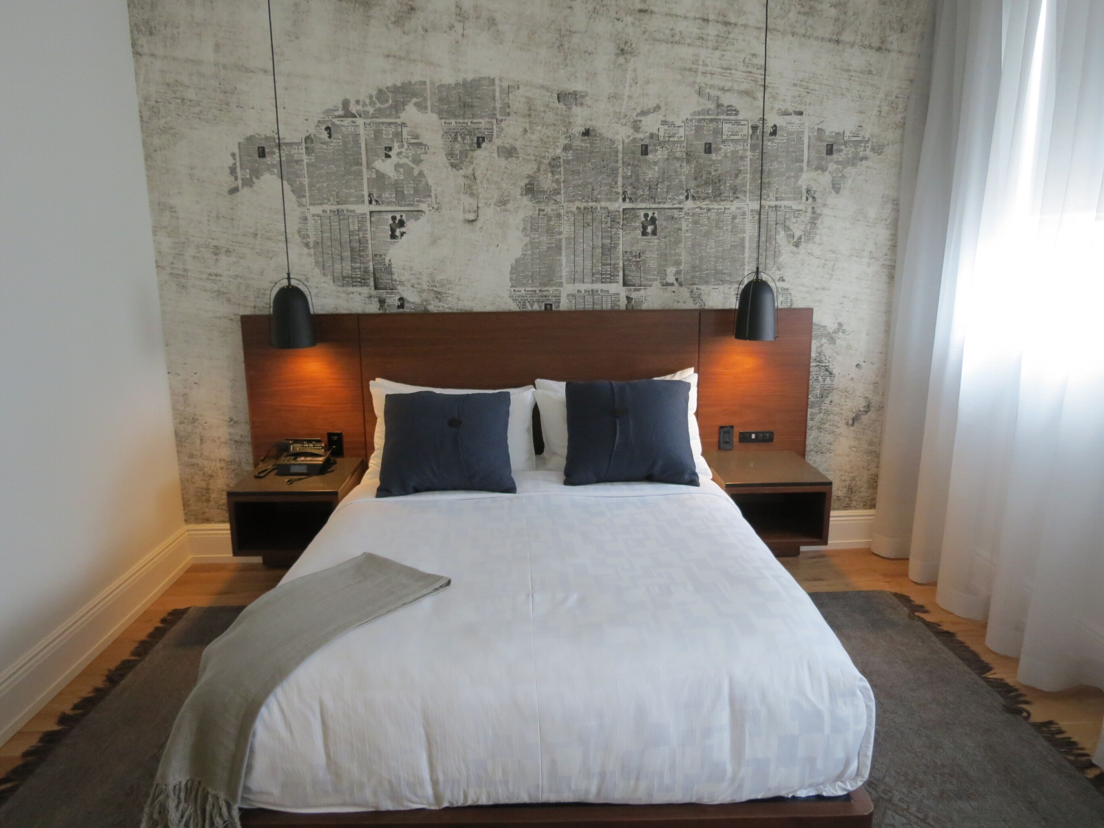 A neatly made bed with a white duvet and two blue pillows, flanked by two bedside tables with lamps, against a wall with a map design.