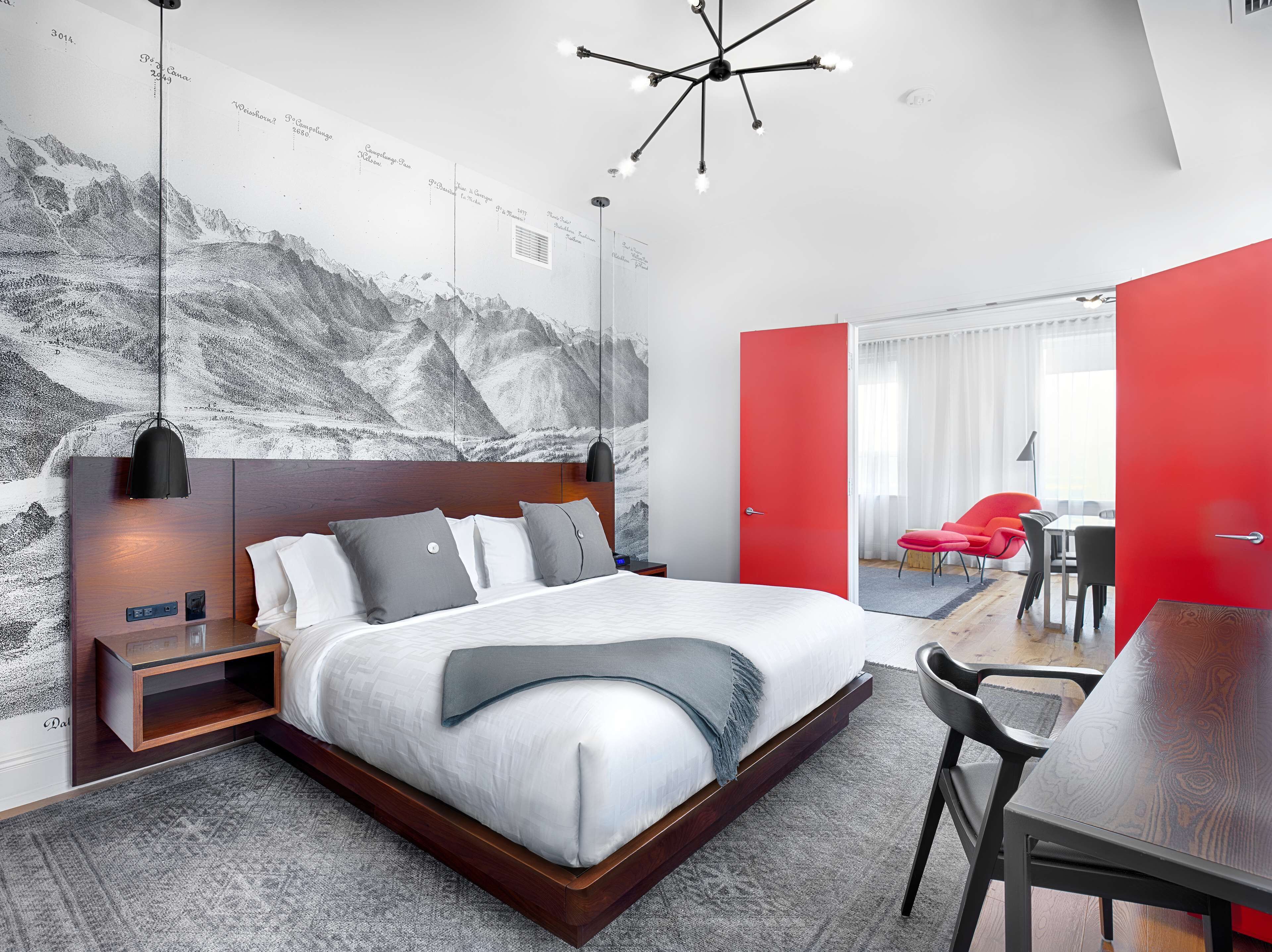 Un dormitorio moderno con una cama grande, una mesita de noche, un escritorio con silla, y una puerta roja que da a un comedor.