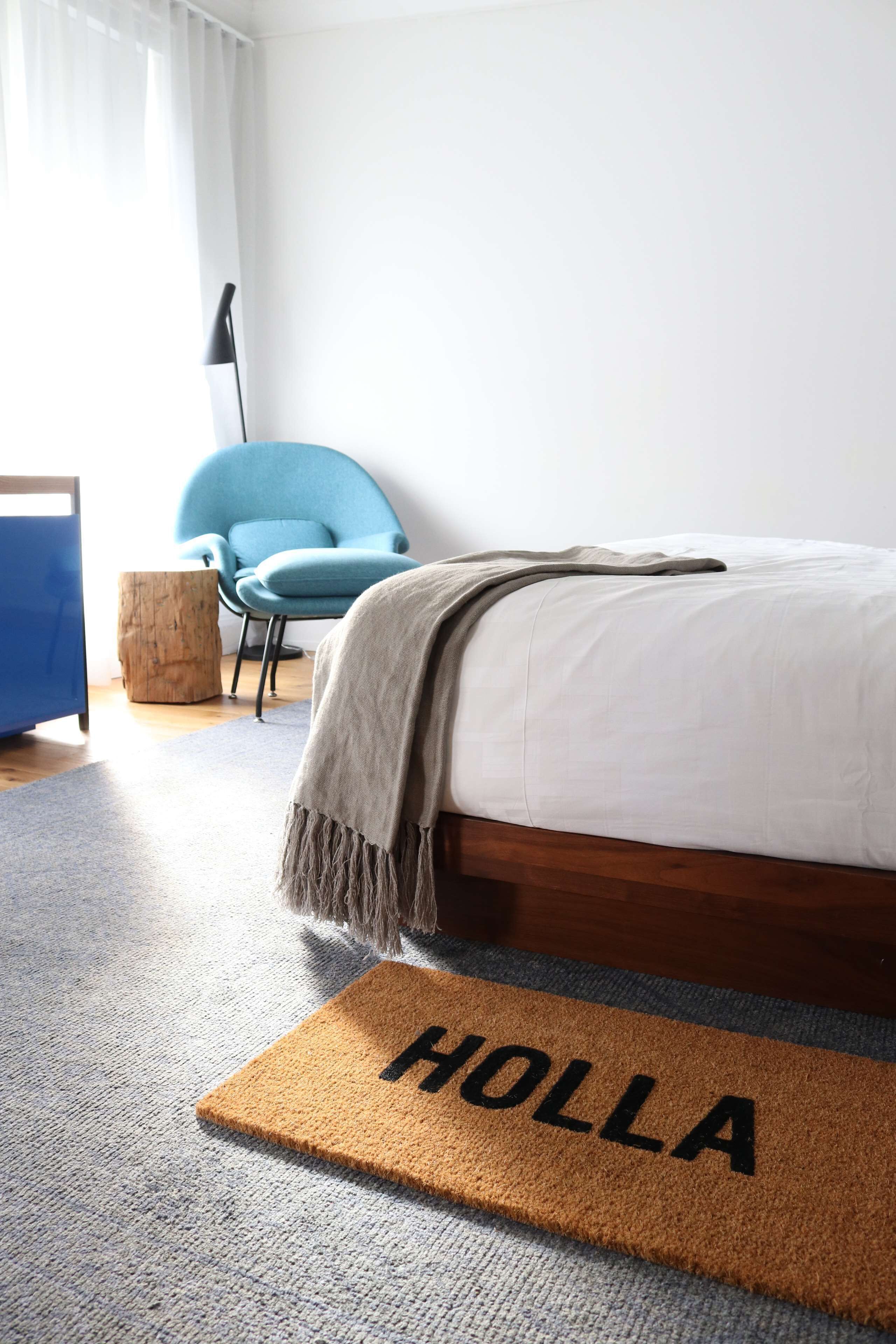 A bedroom with a bed, a blue chair, a wooden stool, and a doormat with "HOLLA" on it.