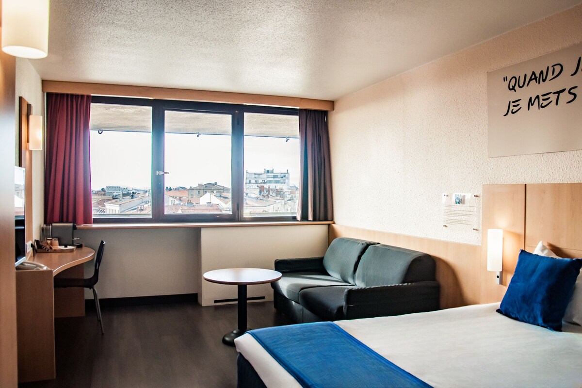 A hotel room with a bed, a desk, a chair, a small table, and a window with curtains.