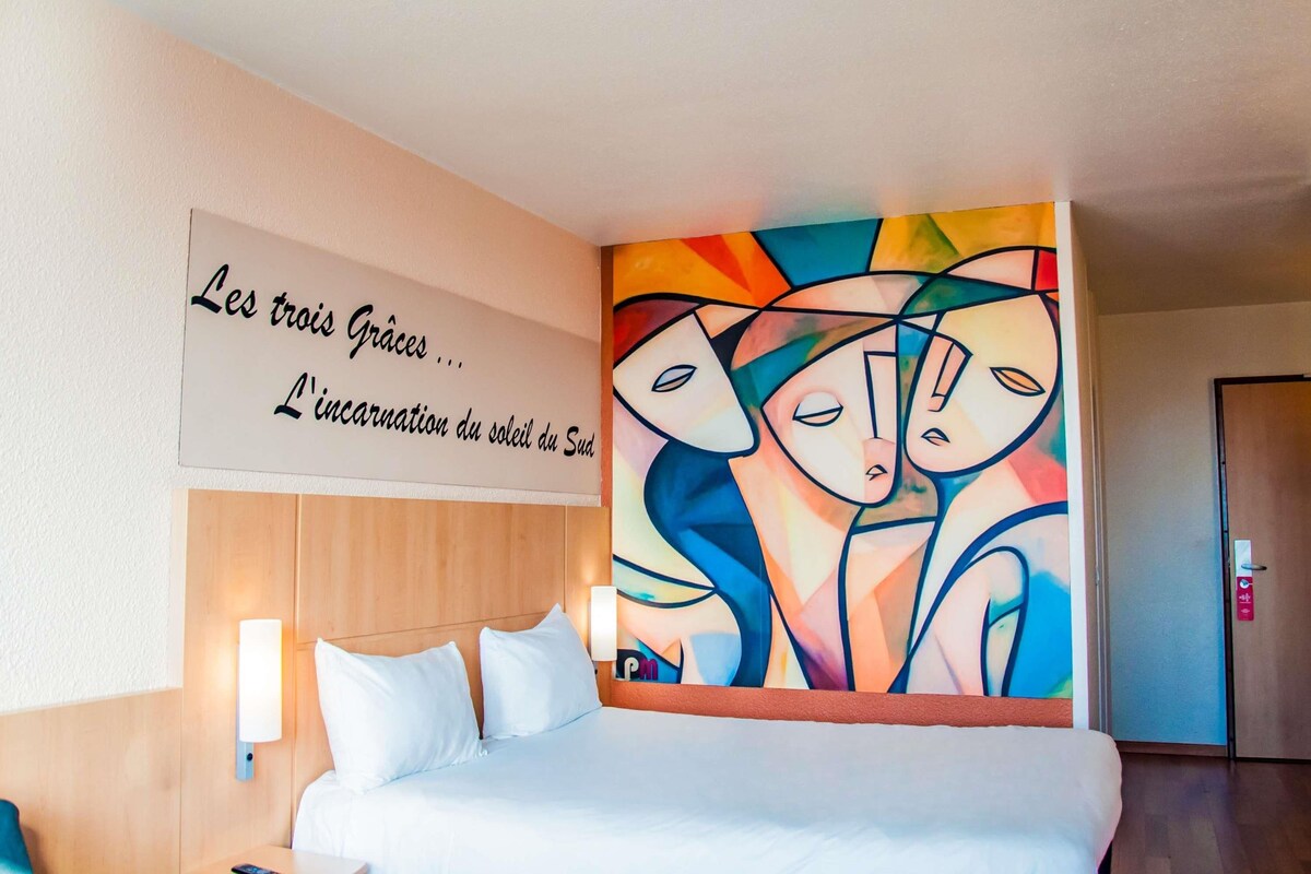 Una habitación de hotel con una cama, un cartel en la pared y un cuadro abstracto de colores vivos.