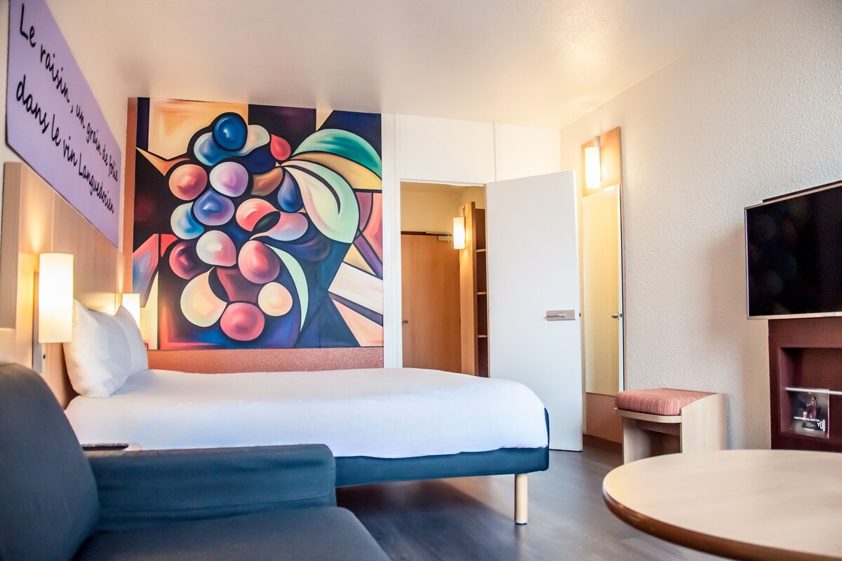 A hotel room with a bed, a painting on the wall, a television, and a small table.