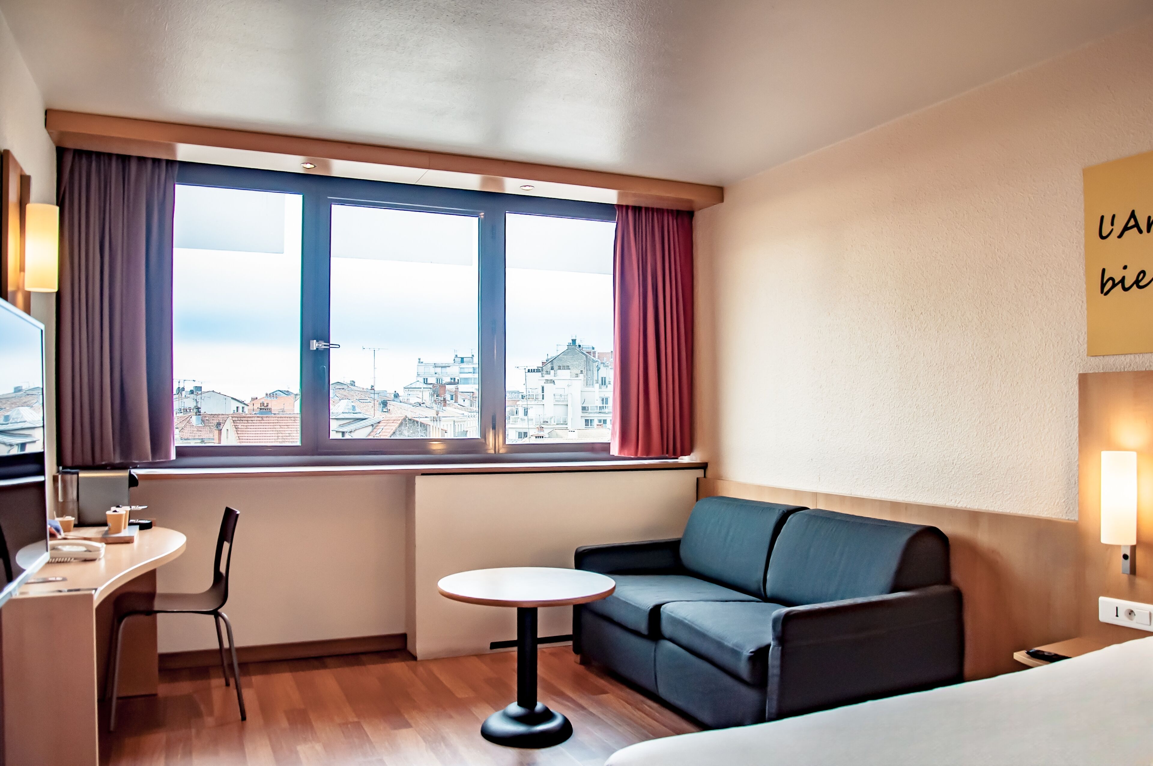 A hotel room with a large window, a sofa, a small table, a desk with a chair, and a bed.