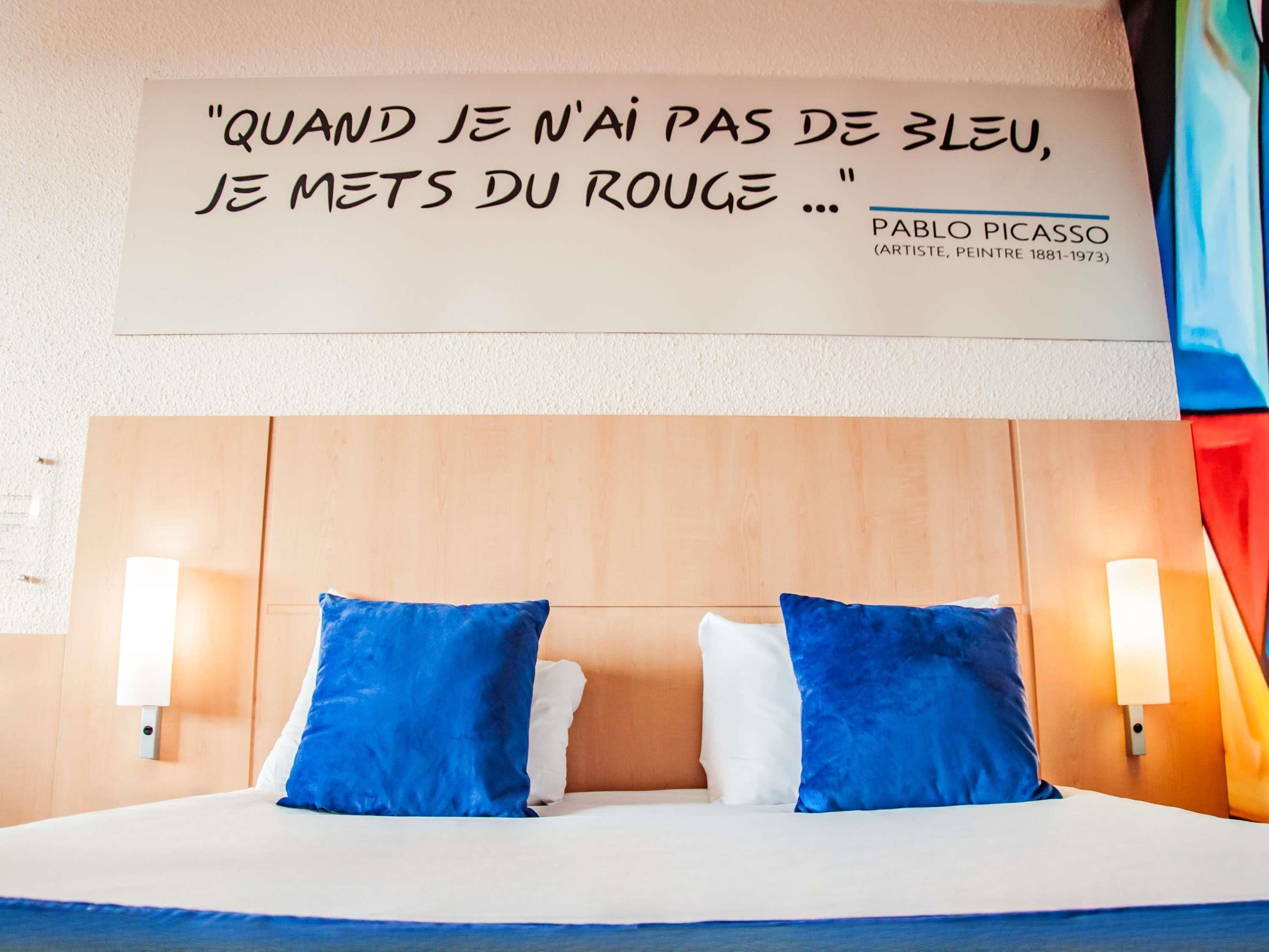 A hotel room with a bed, two blue pillows, a white pillow, and a wall quote by Pablo Picasso.